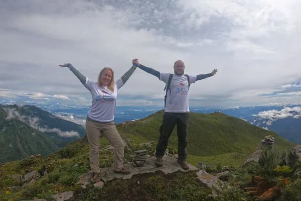 A man and woman wearing matching white "Visit Nepal 2020" t-shirts standing on a mountain peak with arms raised high against a valley backdrop.