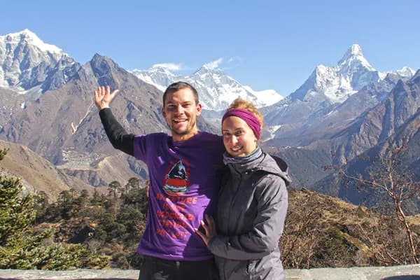 A smiling man in a purple t-shirt and a woman in a grey jacket standing together on a vantage point with Mount Everest