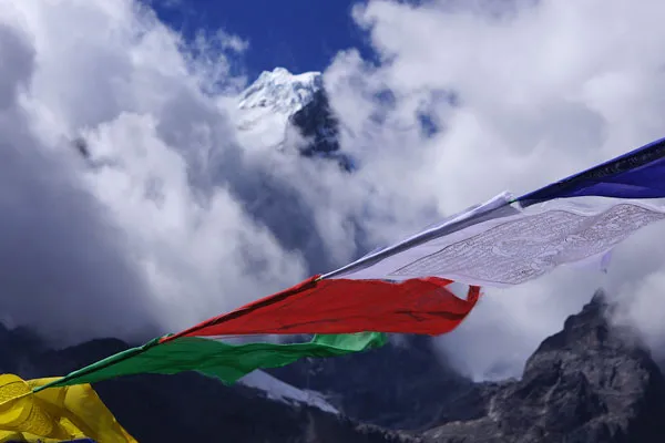 Colorful Buddhist prayer flags blowing in the wind close-up with a dramatic snow-covered mountain peak emerging through clouds.