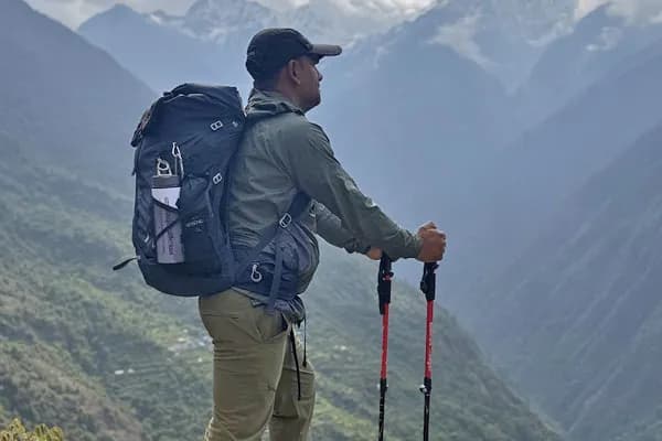 A male trekker in a green jacket and cap holding trekking poles and carrying a blue backpack while looking across a misty mountain valley
