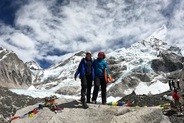 Two women in puffer jackets smiling while standing together on a large rock at Everest Base Camp with Khumbu Glacier in the background