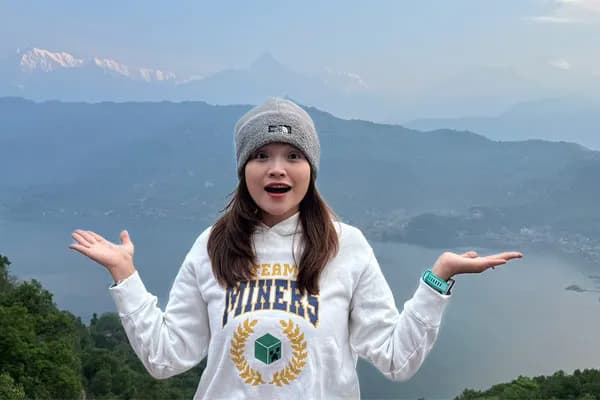 A cheerful young woman in a beanie and white sweatshirt posing with open hands overlooking Phewa Lake and the mountains in Pokhara.