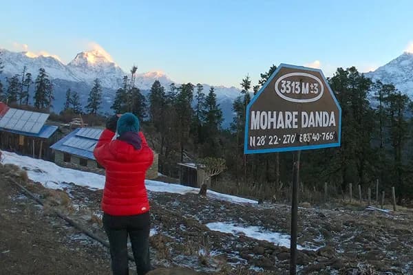A trekker in a red jacket photographing the snow-capped Annapurna mountain range next to a brown Mohare Danda signpost showing 3,313 meters elevation