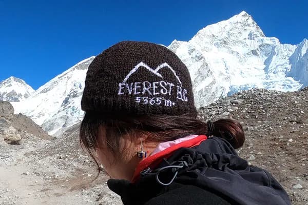 Close-up profile of a trekker wearing a dark brown beanie embroidered with "Everest BC 5364m" against snow-capped peaks