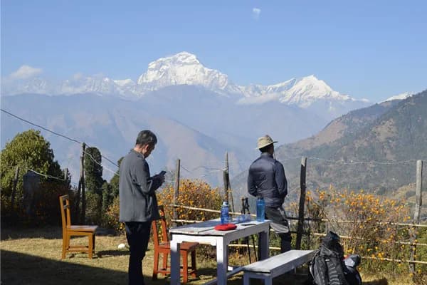 Trekkers resting at an outdoor teahouse table overlooking snow-covered Himalayan peaks in Nepal.
