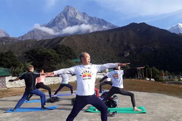 A group of yogis wearing white Everest Yoga Trek shirts practicing Warrior II pose on mats on a stone platform with Himalayan mountains behind them