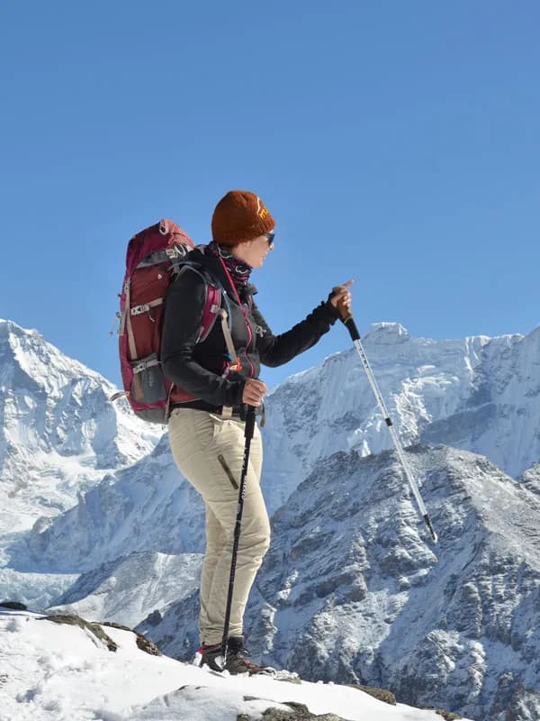 Female trekker with backpack and trekking poles standing on a snowy mountain ridge under a clear blue sky