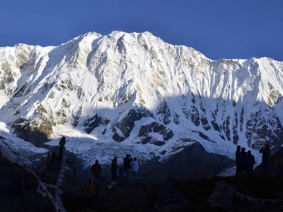 Massive snow-covered face of Mount Annapurna South with silhouettes of trekkers admiring the high-altitude Himalayan peak.