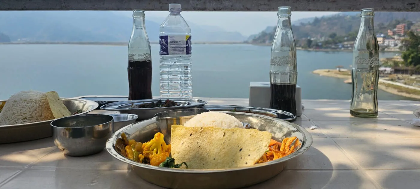 Plates of traditional Dal Bhat served with cold drinks at a scenic lakeside restaurant table