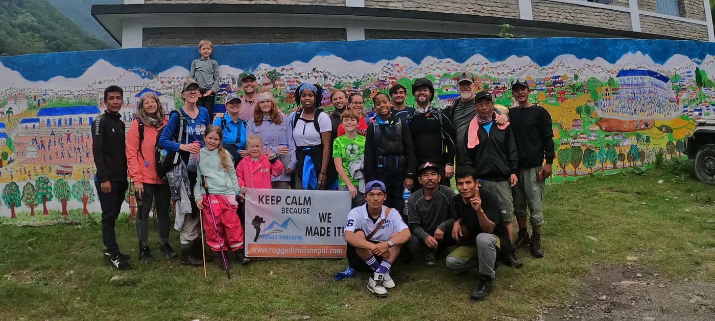 Diverse group of trekkers and local guides holding a Rugged Trails Nepal banner in front of a colorful village wall art.