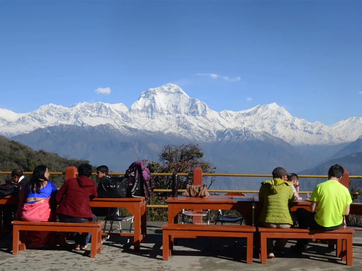 Trekkers relaxing on wooden benches at a mountain teahouse enjoying views of Mount Dhaulagiri and the Annapurna range in Nepal.