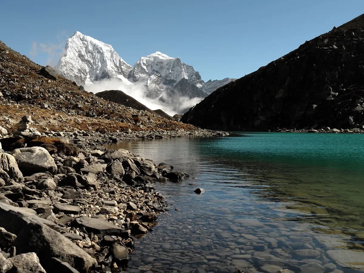 Clear turquoise waters of a Gokyo Lake with rocky shoreline and snow-capped peaks in the background.