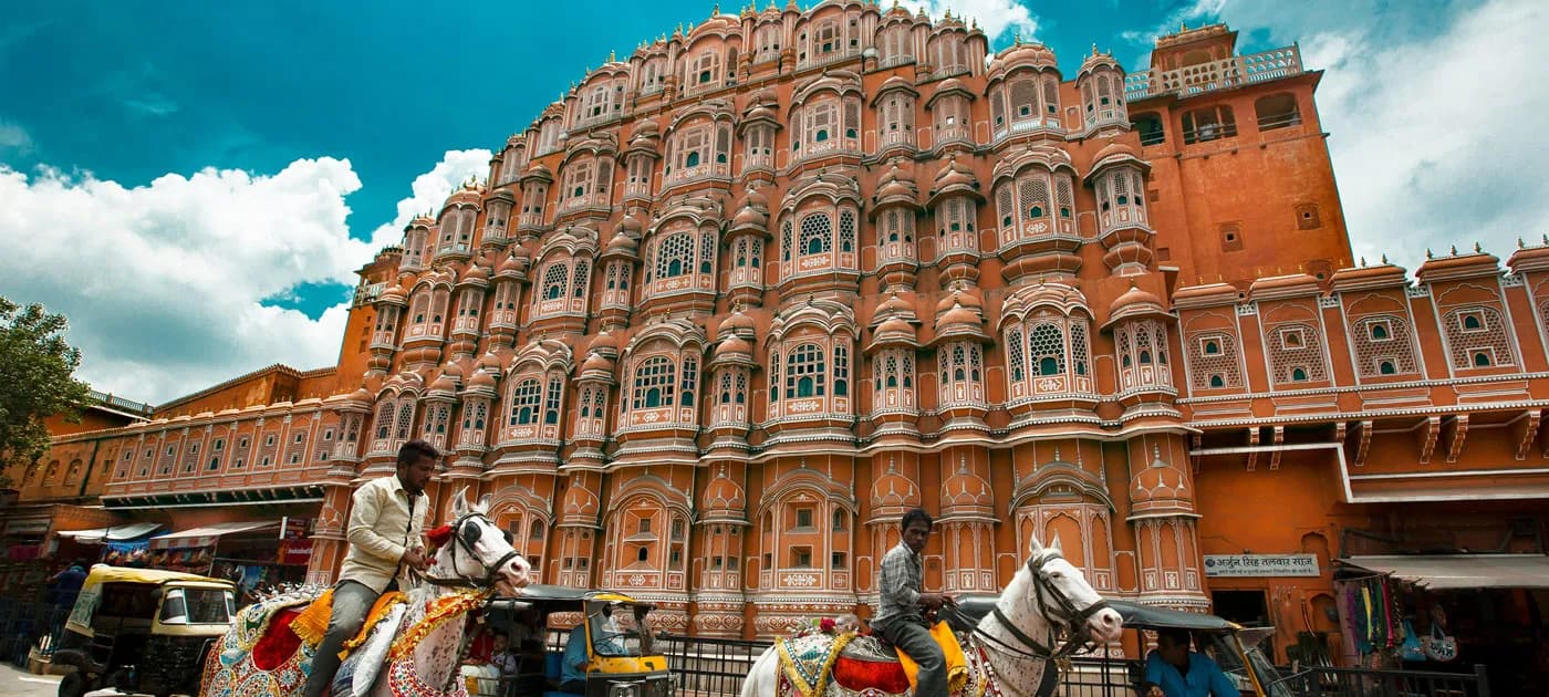 Intricate honeycomb facade of Hawa Mahal in Jaipur with street scenes featuring horses and auto-rickshaws.