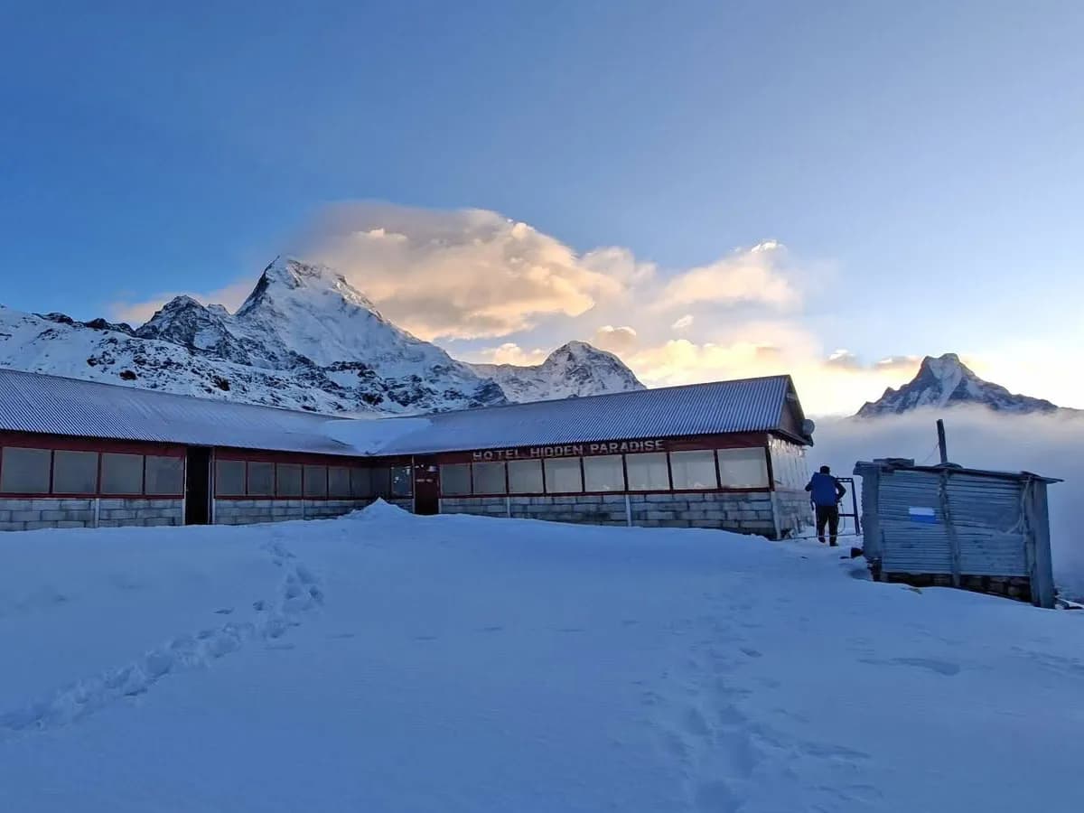 Snow-covered ground leading to Hotel Hidden Paradise teahouse with dramatic Himalayan peaks rising above the clouds.