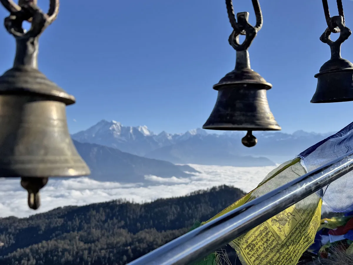 Close-up of traditional Hindu bronze bells and Tibetan prayer flags hanging with a cloud-filled valley and snow-capped Himalayan peaks in the background