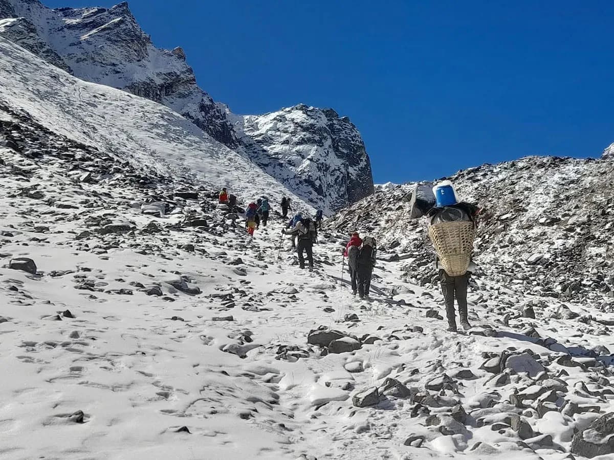 Trekkers and local porters carrying heavy gear ascending a steep, snow-dusted rocky trail towards Ganja La Pass in Nepal.