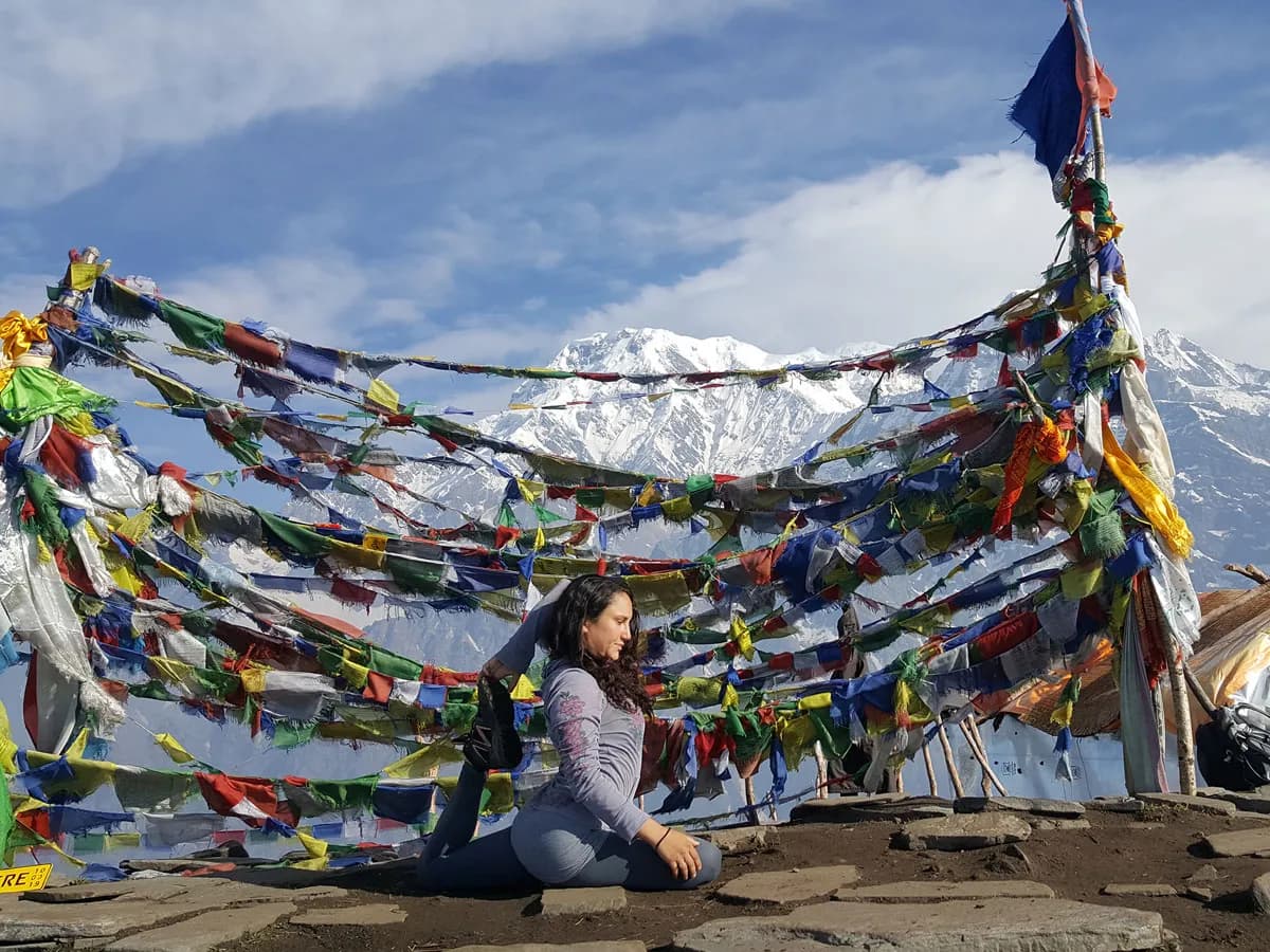 A woman practicing a yoga pose outdoors framed by colourful Buddhist prayer flags with snow-capped Himalayan peaks in the background during the Mardi Himal Yoga Trek.