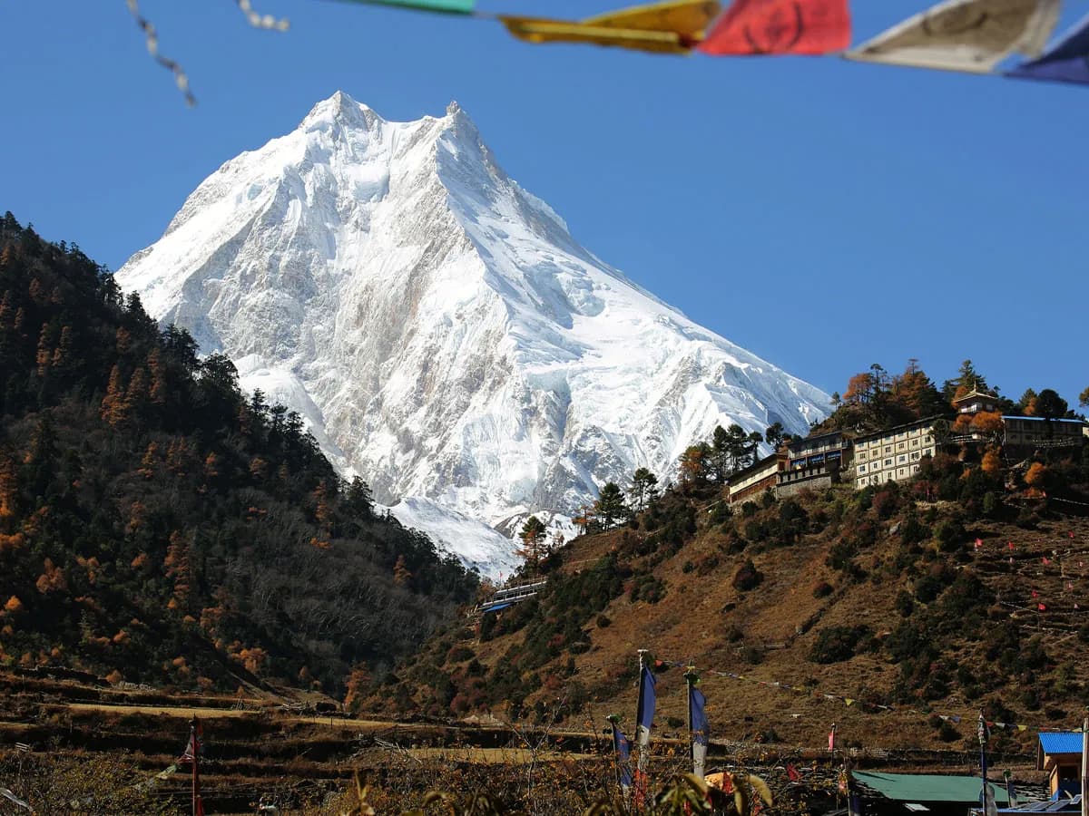 Snow-capped Mount Manaslu peak rising above a monastery village in Nepal, framed by prayer flags against a clear sky