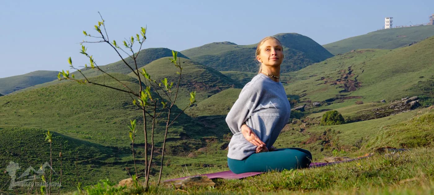 Yogi practicing seated yoga stretch posture on a hill with rolling green mountains under a clear sky