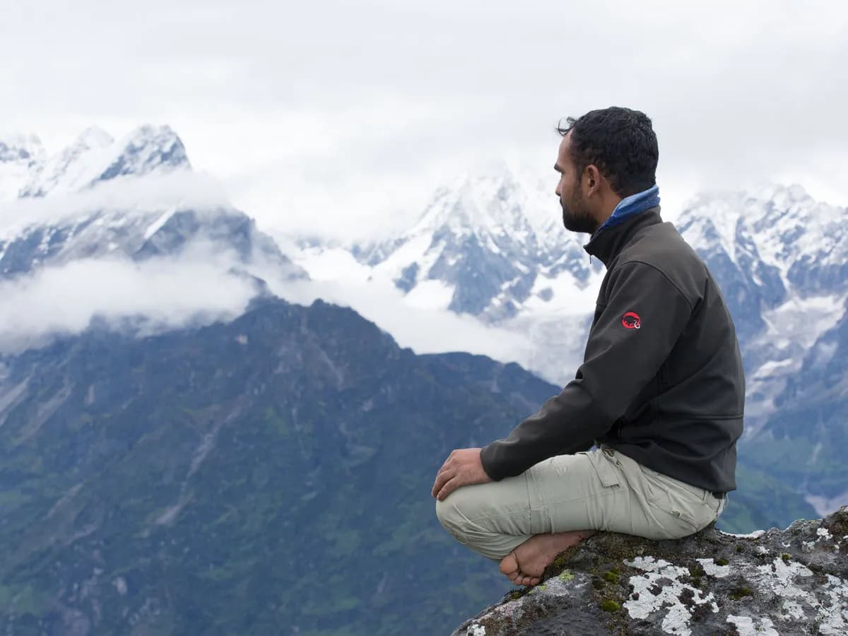 A man sitting in a cross-legged meditation pose on a cliff edge looking out at snow-covered Himalayan peaks in Nepal.