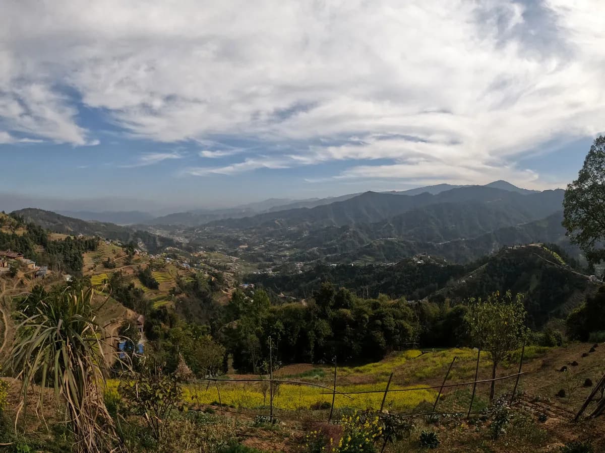 A wide panoramic landscape view of green terraced hills and agricultural valleys under a blue sky with light clouds, taken along the Phulchowki to Namo Buddha trek