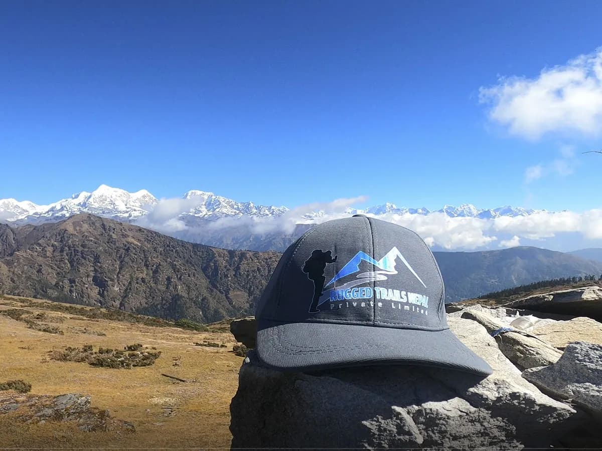 A grey trekking cap resting on a rock ledge overlooking a snow-covered Himalayan mountain range and open ridge pasture under a clear sky