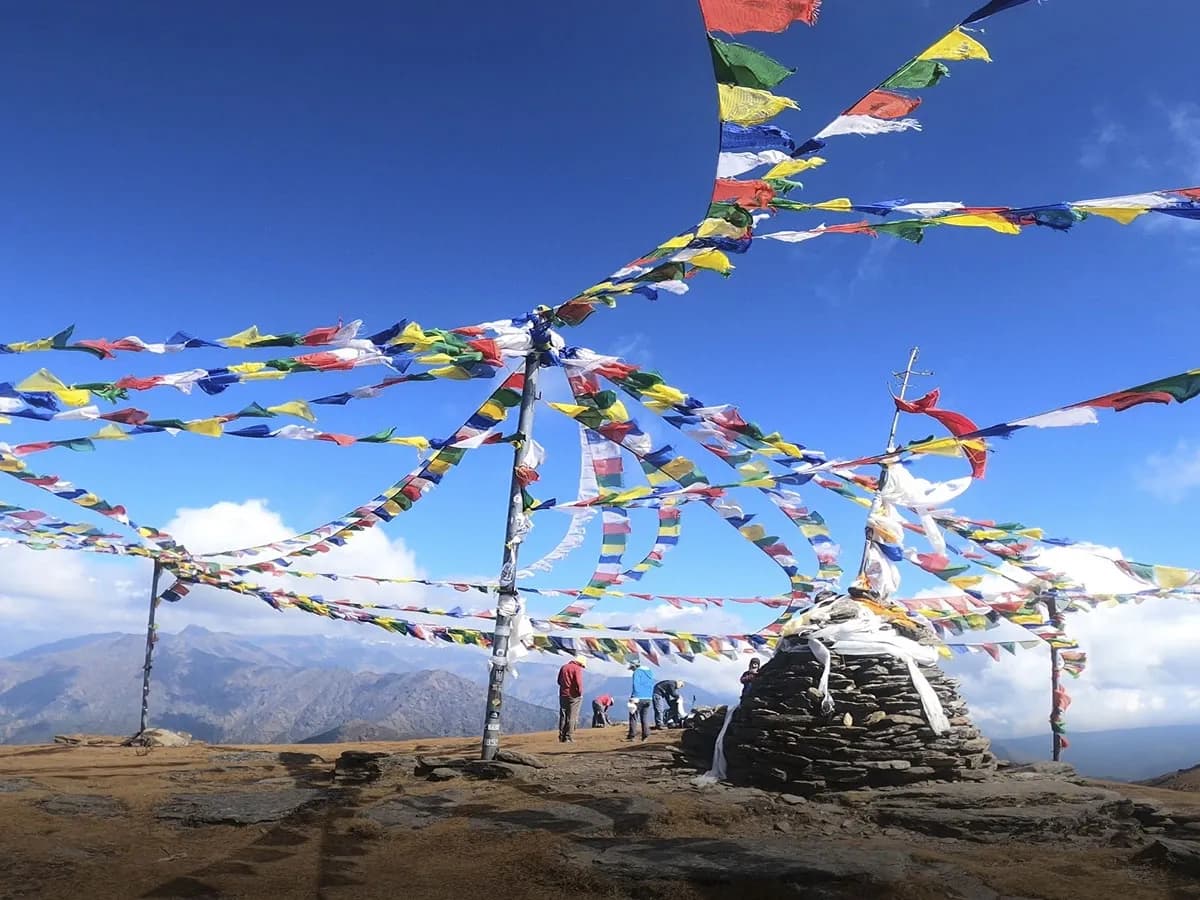 Panoramic view of Buddhist prayer flags radiating from central poles atop a rocky mountain ridge overlooking distant Himalayan peaks