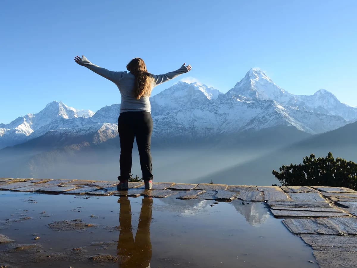 Solo female trekker standing at Poon Hill viewpoint with her reflection in a puddle and Annapurna South in the background