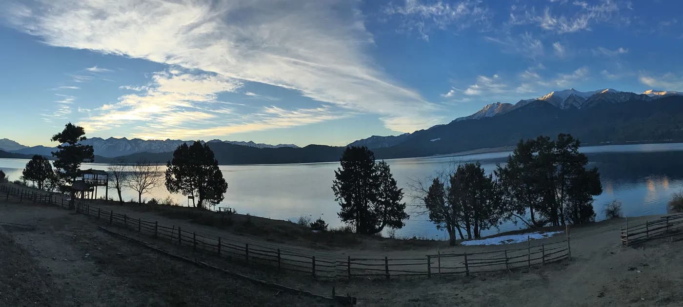 Wide panoramic landscape view of Rara Lake surrounded by dark evergreen trees, a wooden fence, and snow-capped mountain peaks under a blue sky with light clouds.