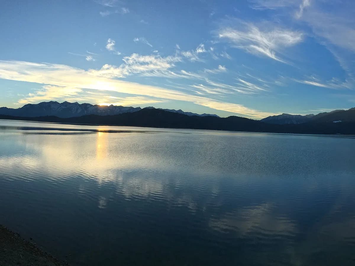 Panoramic wide shot of sunset glowing behind mountain silhouettes across the vast surface of Rara Lake under a light-filled sky