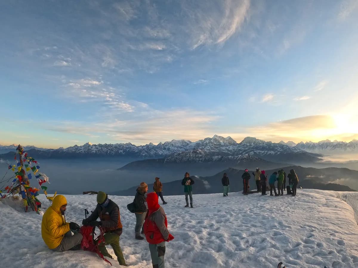 A group of trekkers wearing warm jackets gathering on the snowy summit of Pikey Peak adorned with colorful prayer flags, with a panoramic view of the Himalayan mountain range under a bright morning sun
