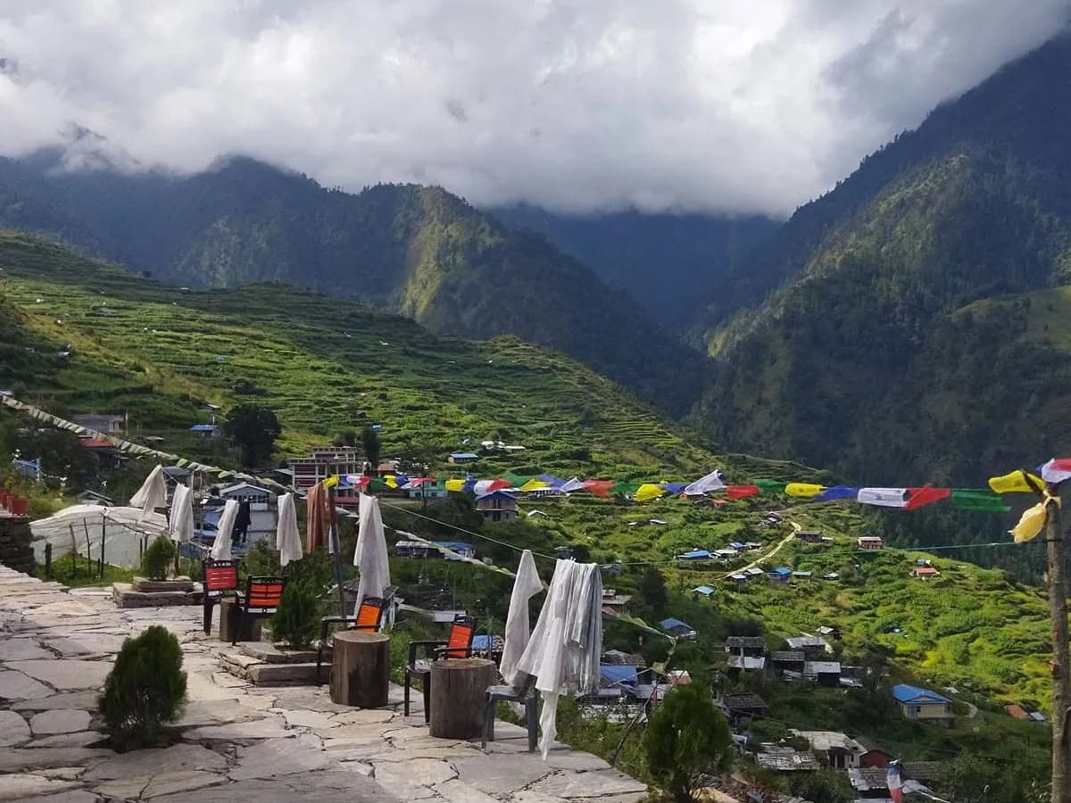 Scenic view of a mountain teahouse patio adorned with prayer flags overlooking lush green terraced fields and a Tamang village in Nepal.