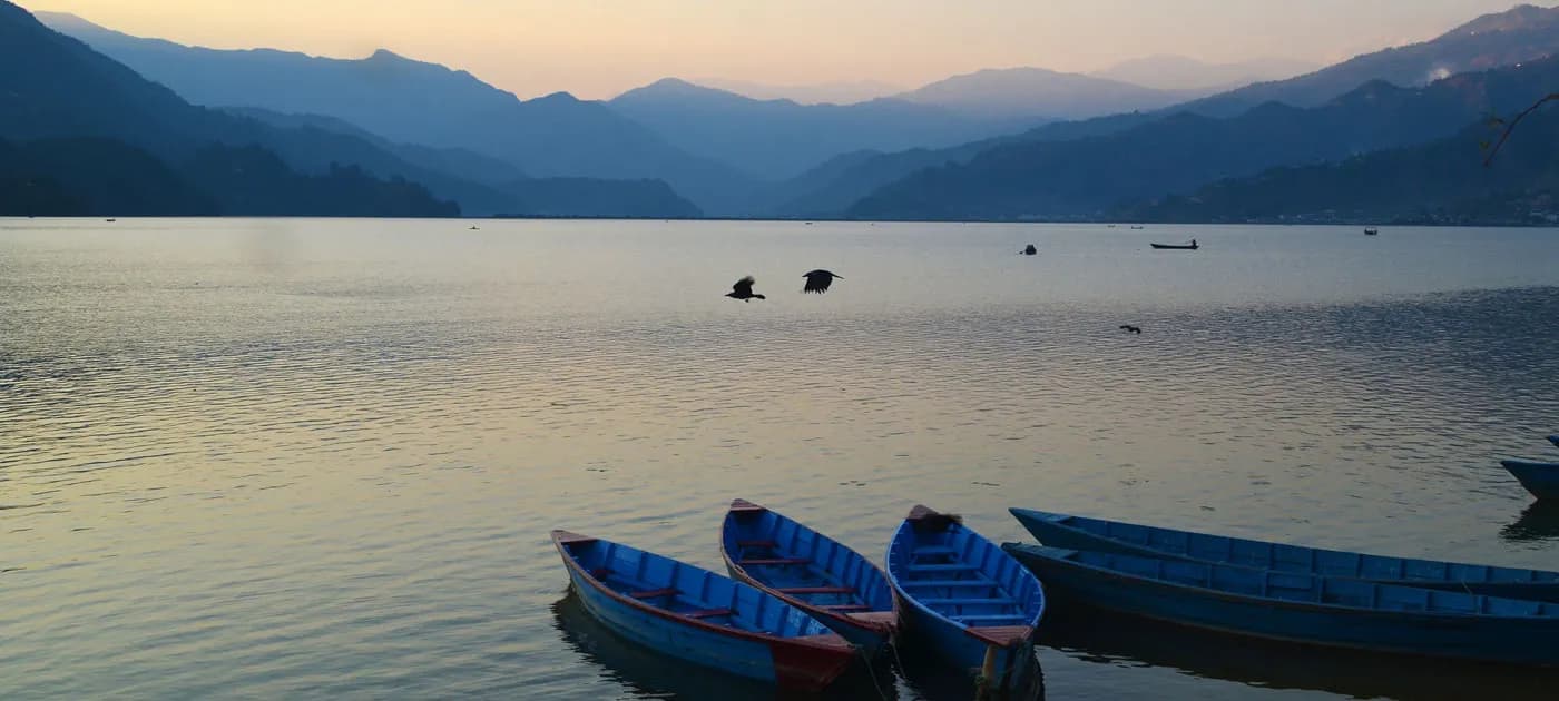 Colorful wooden boats floating on peaceful Phewa Lake with distant mountain hills in Pokhara at sunset.