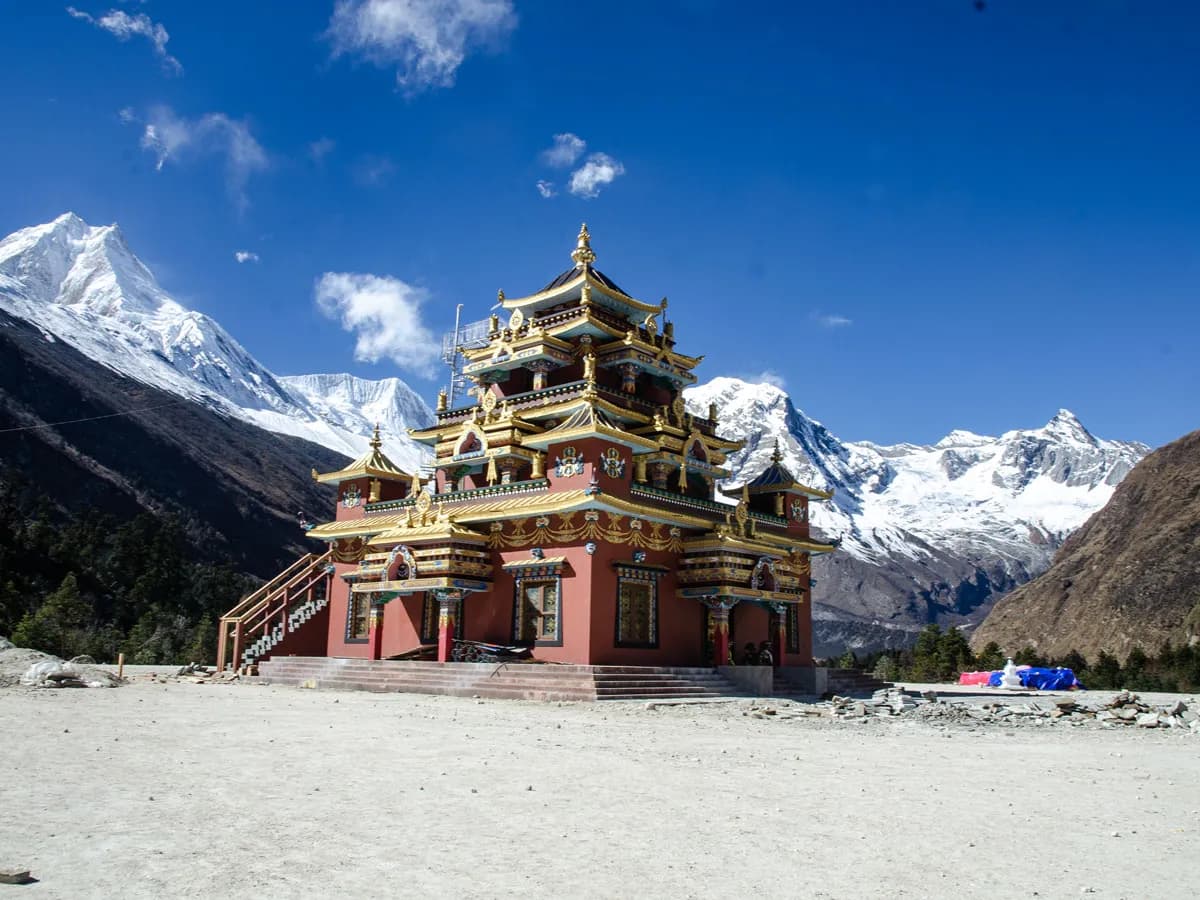 Multi-tiered red and gold Tibetan monastery standing against a bright blue sky and snow-capped Himalayan mountains.