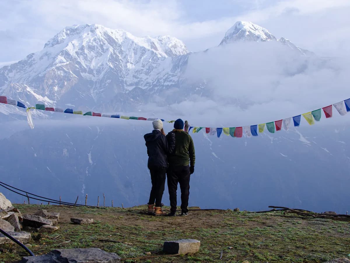 Two trekkers standing together behind colorful prayer flags, gazing at the misty Annapurna mountains on the Mardi Himal Trek.
