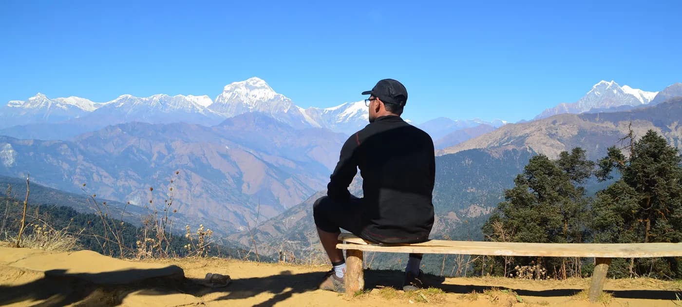 Male trekker resting on a wooden bench overlooking a vast mountain valley and snow-capped Himalayan peaks