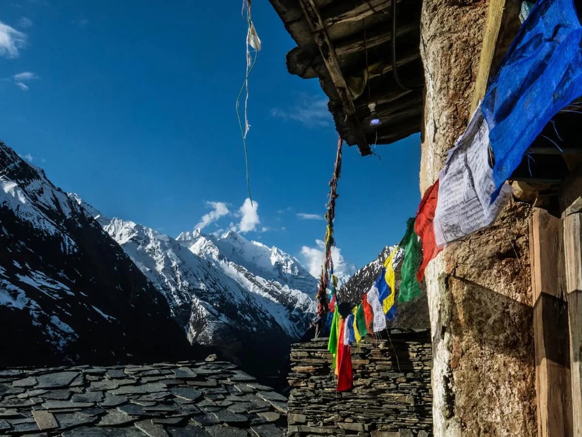 Colorful Tibetan prayer flags fluttering from a stone building corner with snow-capped mountain peaks in the background