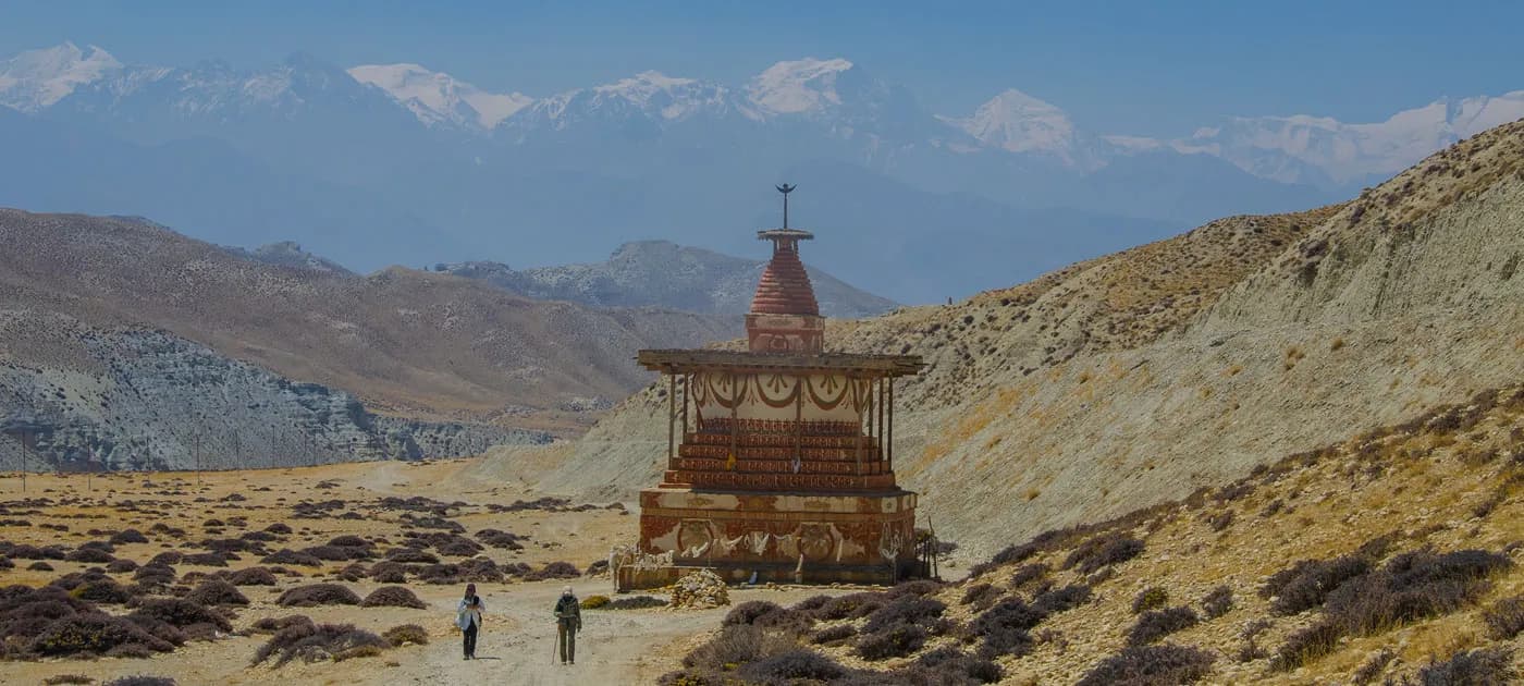 Two trekkers walking past an ancient Buddhist chorten in the arid, high-altitude desert landscape of Upper Mustang.