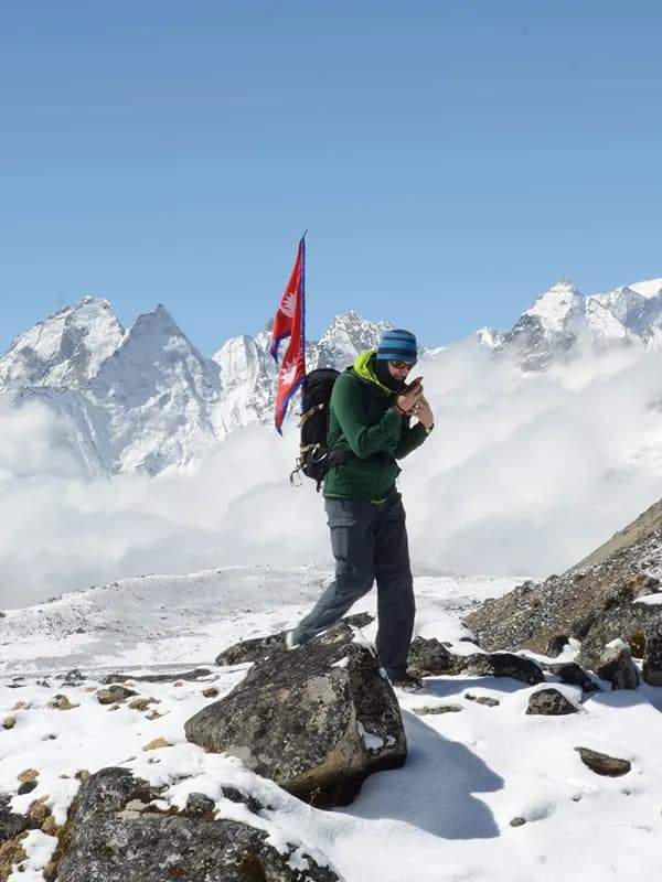 A hiker with a Nepal flag attached to their backpack navigating a rocky, snow-covered ridge high above the clouds