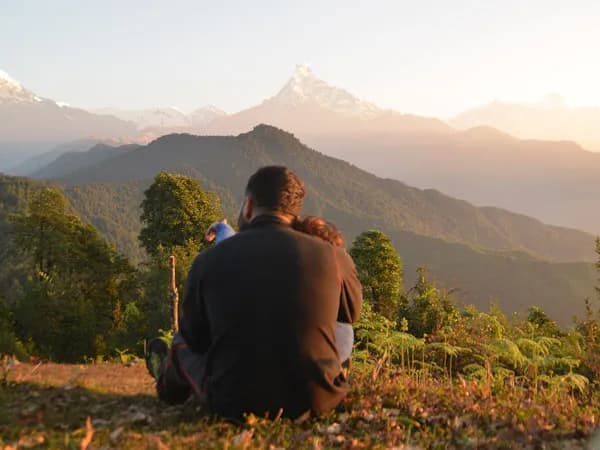 A couple sitting together on a grassy hill from behind, watching the golden morning sunlight illuminate the peak of Machhapuchhre (Fishtail) mountain and the Annapurna range