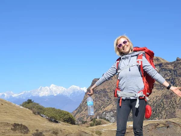 A smiling woman wearing a backpack and sunglasses standing with open arms on a grassy mountain ridge with snowy Himalayan peaks in the background