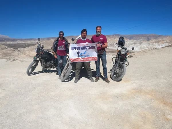 Three adventure riders standing with motorcycles on an arid dirt road in Upper Mustang, holding up a banner that reads 'Here We At Upper Mustang