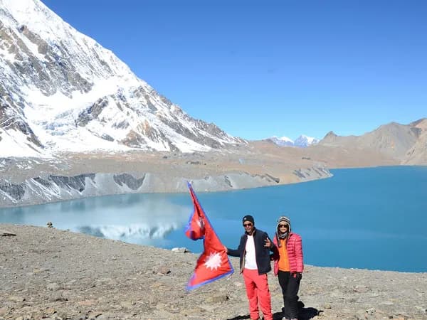 Two trekkers standing by the turquoise waters of Tilicho Lake holding the national flag of Nepal, with snow-covered Himalayan peaks reflecting on the water under a clear blue sky.