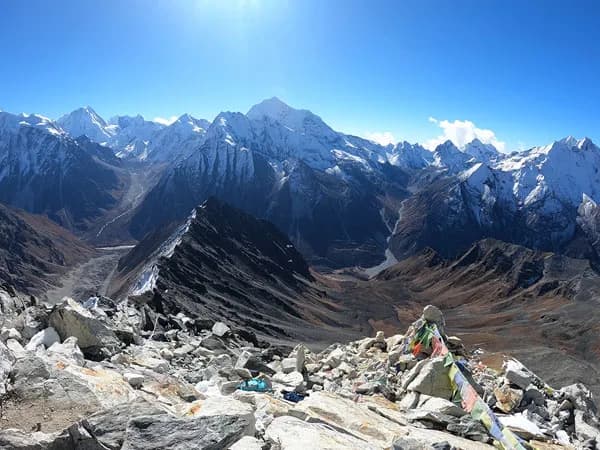 A high-altitude mountain summit adorned with colorful Buddhist prayer flags, overlooking rugged rock ridges and snow-capped Himalayan ranges under a clear blue sky