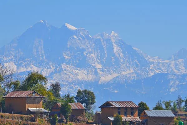 Traditional clay and brick village homes set against a clear blue sky and towering snow-covered Himalayan peaks.