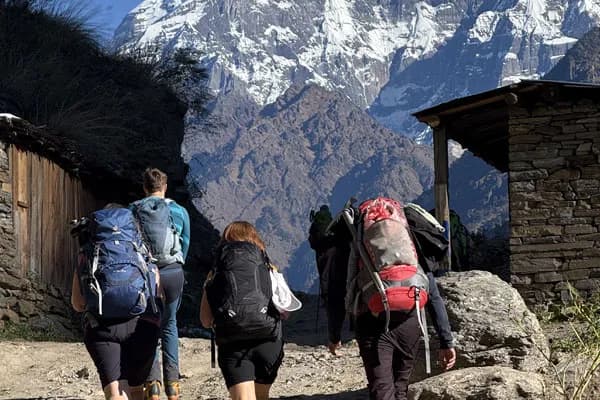 A group of trekkers wearing heavy backpacks walking up a gravel path past traditional stone houses toward towering snow-capped mountains