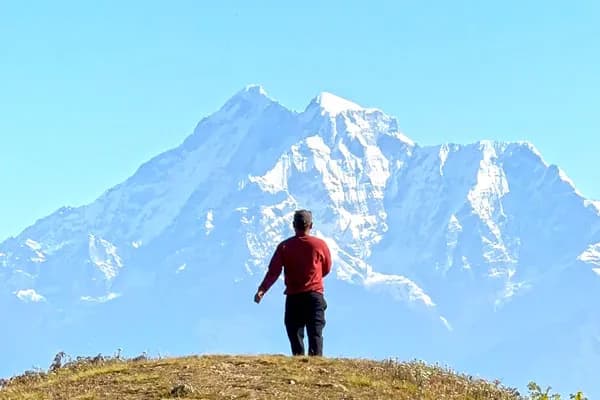 A lone person in a red sweater standing on a grassy hill facing a massive snow-covered mountain peak