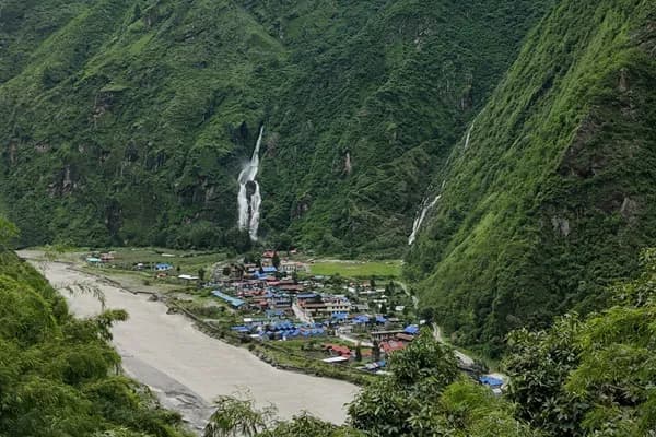 A lush green mountain valley in Nepal with tall waterfalls cascading down steep hillsides past a riverside village during the monsoon season