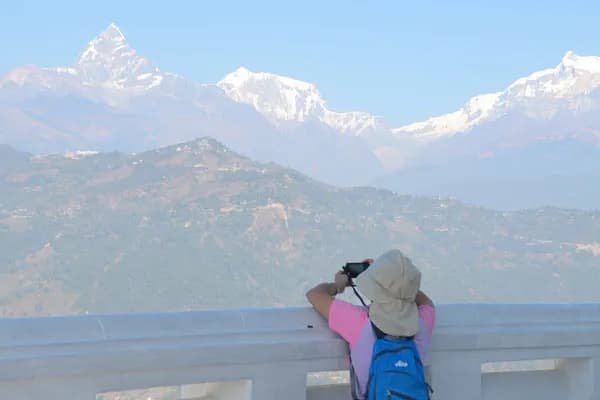 A traveler wearing a sun hat and blue backpack taking a photo of Machhapuchhre (Fishtail Mountain) from a white terrace viewpoint in Pokhara.