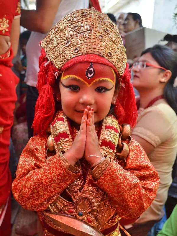 Young girl dressed as the Living Goddess Kumari in traditional red attire and golden crown offering Namaste greeting